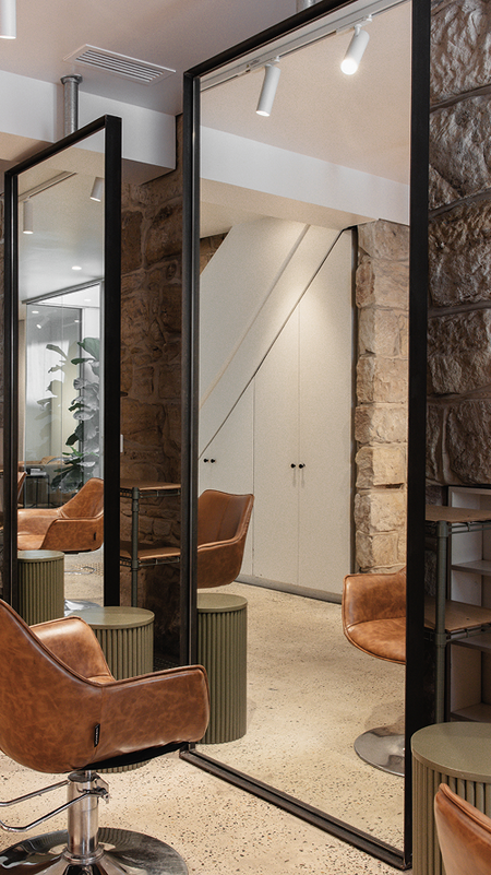 A **brown leather swivel chair** sits elegantly in front of a sleek mirror, framed by warm **sandstone walls**, with an **olive-toned side table** beside it—creating a refined and inviting space at Neighbourhood Salon, Darlinghurst.