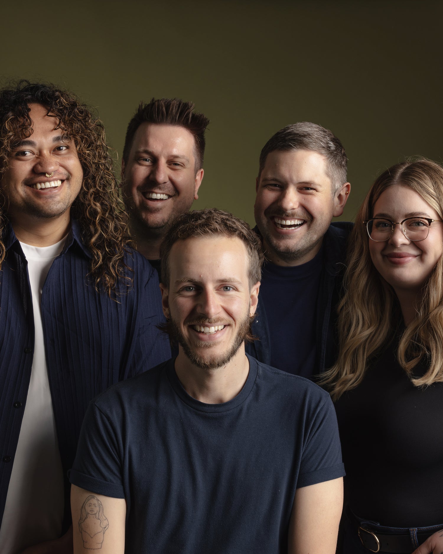A group photo of the **Neighbourhood Salon** team in **Darlinghurst, Sydney, near Surry Hills**, standing together against an **olive green background**. Their diverse styles and friendly energy shine through, reflecting the salon’s warm and inviting atmosphere.
