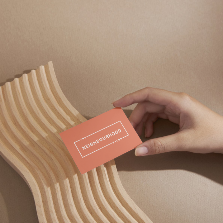 A hand gracefully holds an **orange Neighbourhood Salon gift card** against a warm sandstone-coloured background, highlighting its sleek design and the perfect gift for luxury hair care experiences in Darlinghurst, Sydney.