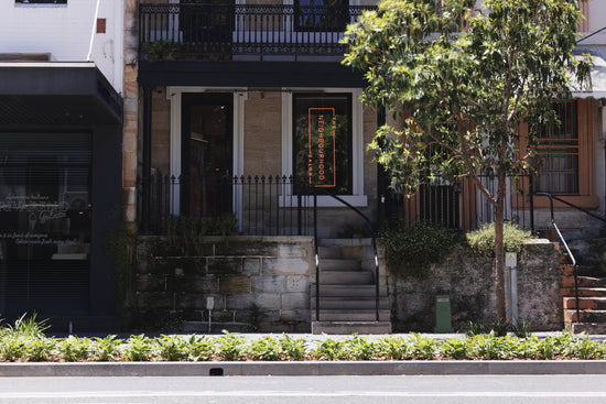 A photo of the **Neighbourhood Salon** exterior in **Darlinghurst, Sydney, near Surry Hills**, showcasing its charming **three-story terrace** facade. A glowing **neon sign** adds a modern touch, while a **lush tree out front** frames the entrance, creating a welcoming atmosphere.